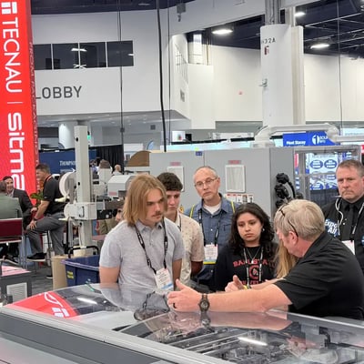 At the PRINTING United trade show, an exhibitor from Tecnau | sitma explains industrial printing equipment to a focused group of visitors gathered around a glass display; the mood is engaged and informative.