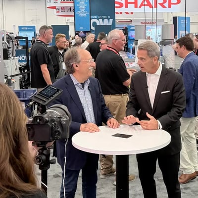 At a crowded PRINTING United technology trade show, two suited men discuss a product across a round white table as a camera films them; lively, professional atmosphere.