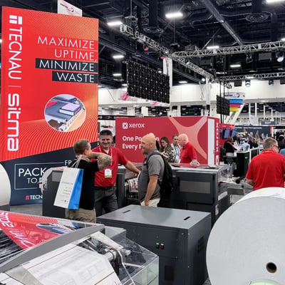 At a busy PRINTING United trade show, attendees talk beside industrial presses and a huge paper roll; bold red displays promote print efficiency.
