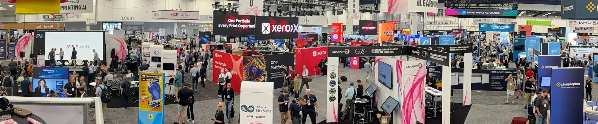 A crowded PRINTING United technology trade show fills a convention hall with colorful company booths, product displays, and visitors networking in a lively atmosphere.