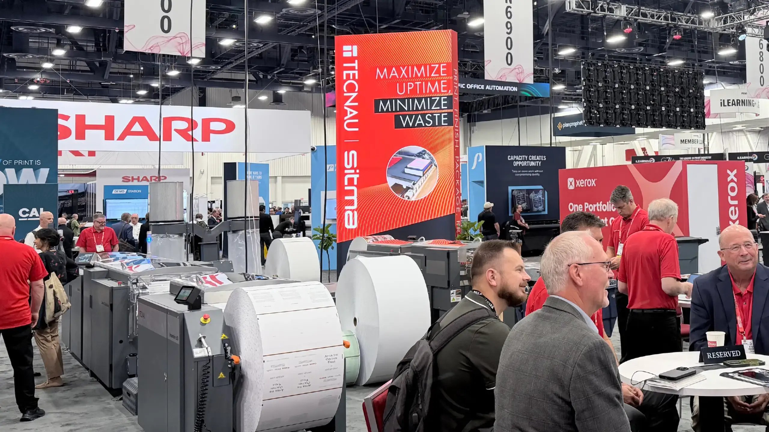 A bustling PRINTING United trade show features large-format printers, Technau | sitma booth, bold signs, and attendees networking under bright lights.